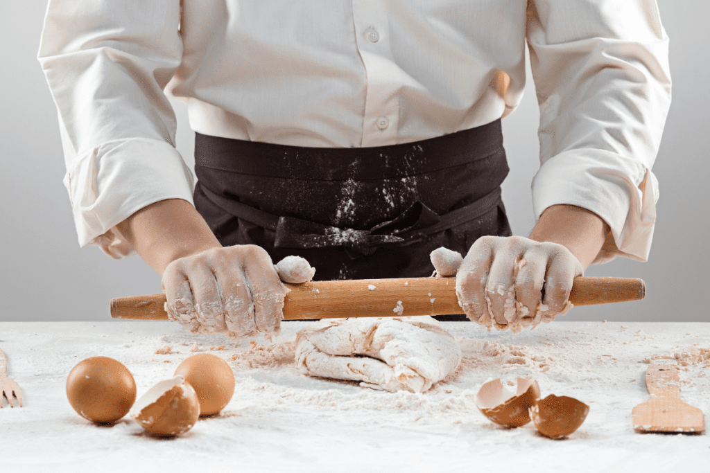 A person rolling dough on a table surrounded by eggs and various baking ingredients.