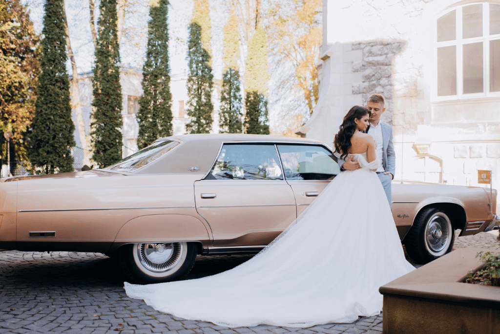 Bride and groom standing together in front of a vintage classic car, smiling and dressed in wedding attire.