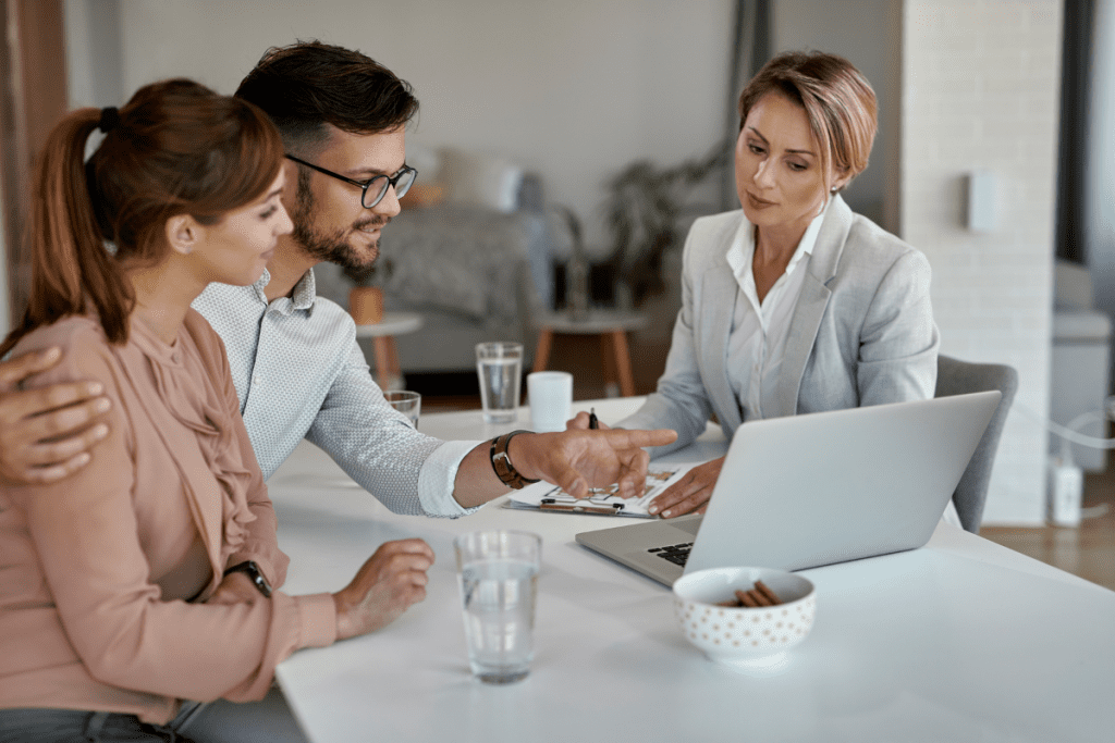 A man and woman sit at a table, focused on a laptop, engaged in conversation or collaboration.