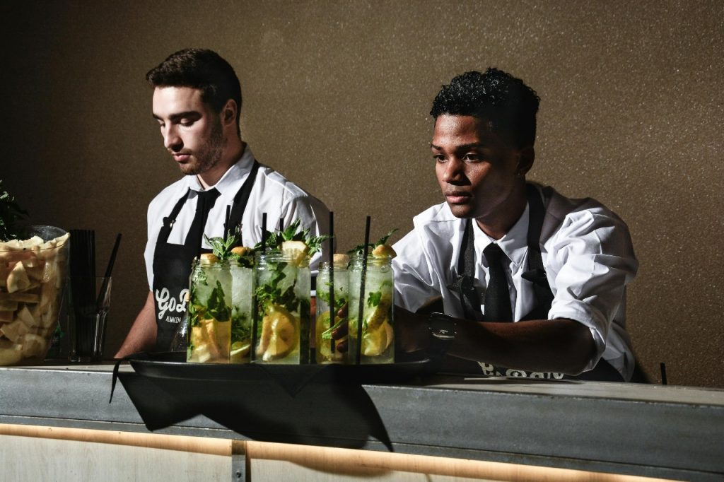 Two men in black shirts and ties standing behind a bar, engaging with customers.